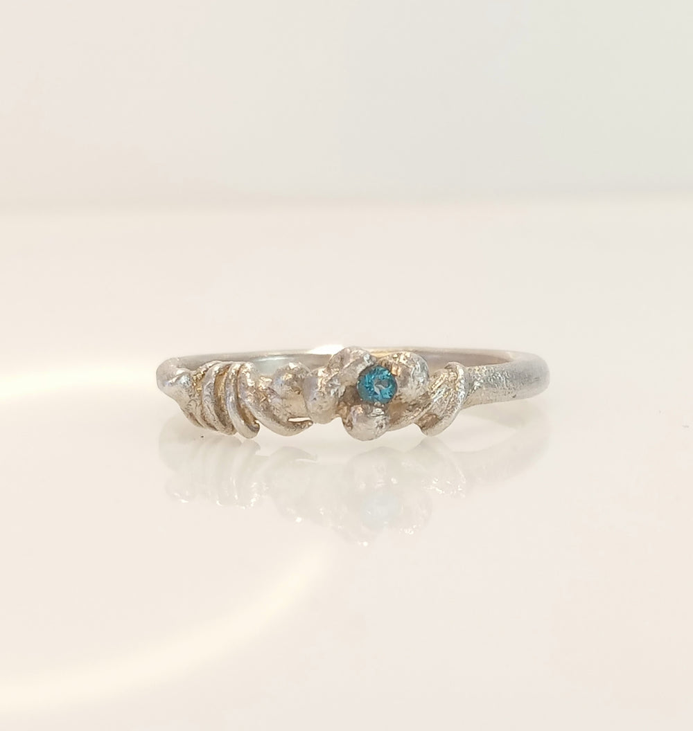 Unser handgefertigter Paraiba Topas Ring in Köln Ehrenfeld ist ein Meisterwerk mit einem tiefblauen 2 mm großen Stein, der das Mittelmeer im Sommer widerspiegelt. Das Band besteht aus 2 mm rundem Silber, und der Stein ist kunstvoll in verschmolzenem Silberdraht gefasst.   Das Band ist aus 2mm rundem Silber gefertigt, der Stein ist in einem Nest aus verschmolzenem Silberdraht gefangen.  Der Edelstein Topas ist ein sanfter, einfühlsamer Kristall, der beruhigt, heilt und auflädt.