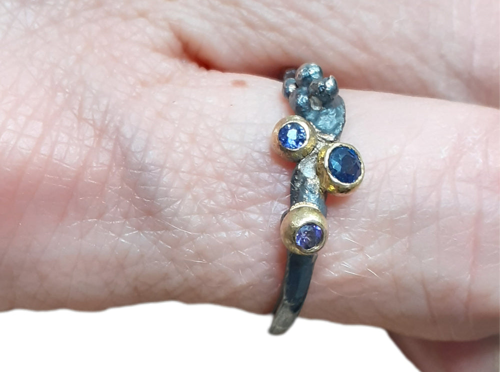 Dieser Ring wurde oxidiert, um den Kontrast zwischen Gold und Silber zu verstärken. Die Steine sind in einer Palette von dunkelblau, lila und lavendel gehalten. Der 3 mm große Stein ist ein Saphir, während die beiden anderen Steine Iolith sind. Wie immer ist das Gold und Silber recycelt.