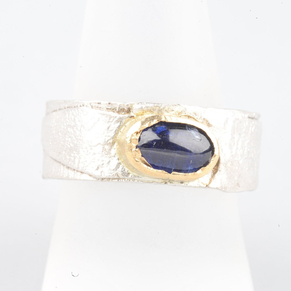 
                  
                    Dieser Ring hat ein schönes Gewicht. Das Band ist 8 mm breit und 2 mm dick. Die Oberfläche wurde retikuliert (ich bringe die Oberfläche des Silbers zum Schmelzpunkt und lasse sie abkühlen. Dies wird mehrmals wiederholt, bis ich mit der Oberflächenstruktur zufrieden bin), während die Innenseite des Bandes glatt und poliert ist. Der Cyanit hat ein wunderschönes tiefes Blau. Die goldene Fassung bringt dies schön zur Geltung.
                  
                