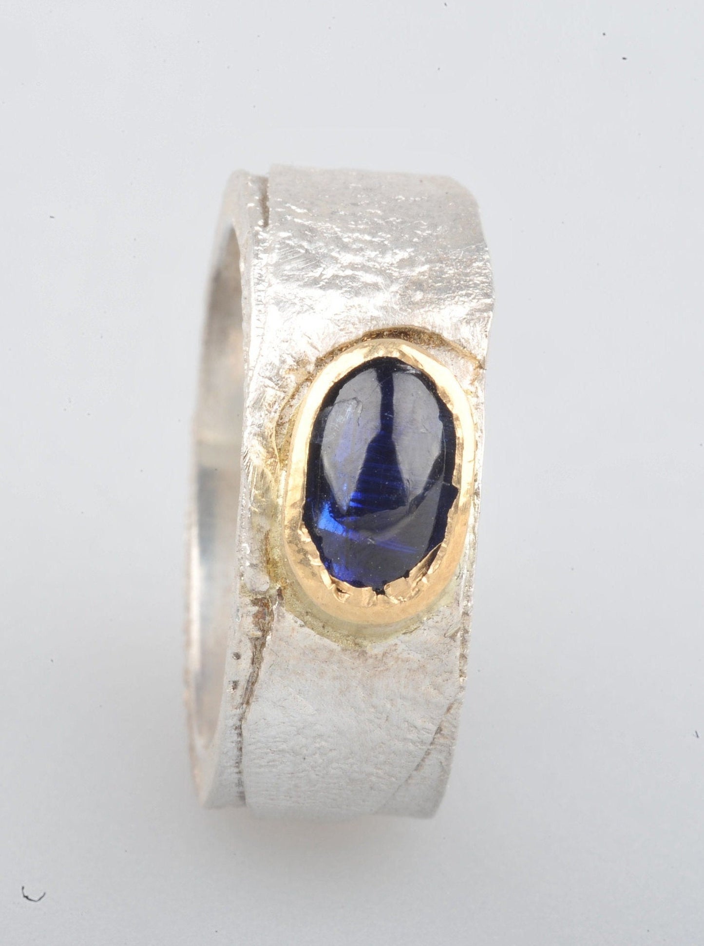 
                  
                    Dieser Ring hat ein schönes Gewicht. Das Band ist 8 mm breit und 2 mm dick. Die Oberfläche wurde retikuliert (ich bringe die Oberfläche des Silbers zum Schmelzpunkt und lasse sie abkühlen. Dies wird mehrmals wiederholt, bis ich mit der Oberflächenstruktur zufrieden bin), während die Innenseite des Bandes glatt und poliert ist. Der Cyanit hat ein wunderschönes tiefes Blau.
                  
                